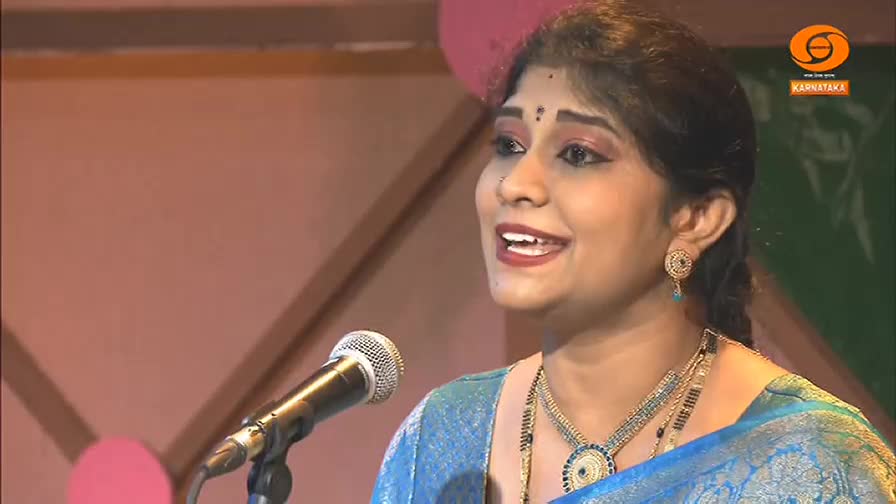 A woman in a blue sari speaks into a microphone, her face lit with a warm smile. Behind her, a logo for DD Chandana, a Kannada-language television channel in...