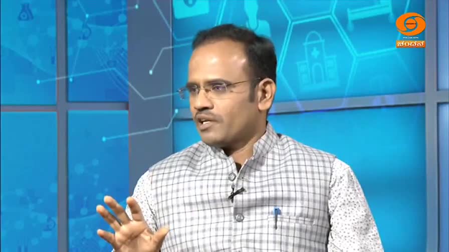 The man, wearing glasses and a checkered vest, gestures with his right hand as he speaks. A DD Chandana logo is visible in the corner, and the backdrop is a ...