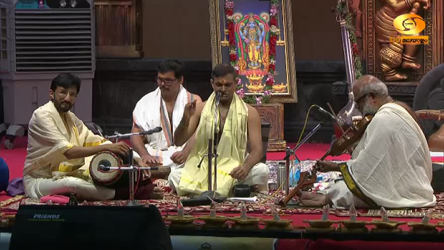 The vocalist, draped in a yellow shawl, gestures while singing into a microphone. A musician with a white beard draws his bow across a violin, adding to the ...