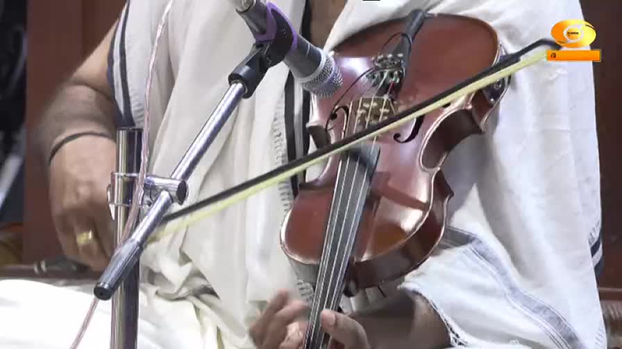 A musician, dressed in white, draws the bow across the violin's strings. The instrument is amplified by a microphone, likely for a broadcast on DD Malayalam.