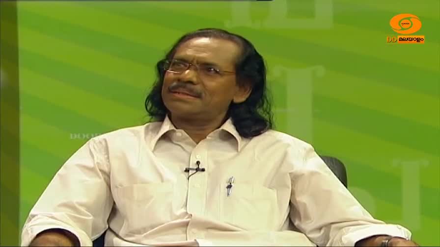 A man in a white shirt sits, looking slightly off-camera, with a DD Malayalam logo visible in the corner. He's in a studio setting, against a green striped b...