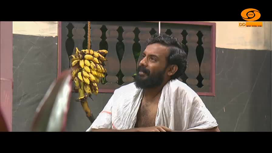 A man with a beard, wearing a white cloth, is speaking, his gaze directed upwards. A bunch of yellow bananas hangs from a rope nearby.