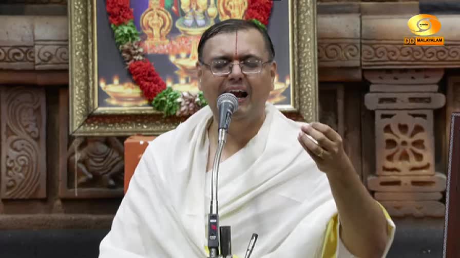 A man in glasses, wearing a white shawl, is singing into a microphone. His right hand is raised, and behind him is a framed image, all broadcast on DD Malaya...