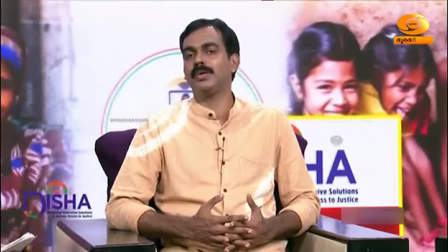A man in a light-colored shirt sits in a studio, speaking with his hands. Behind him, a screen displays images of children and a logo for "Disha."