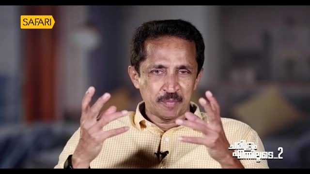 The man on Safari TV gestures with both hands, his fingers splayed as he speaks. He's wearing a yellow patterned shirt, and the logo is visible in the corner.