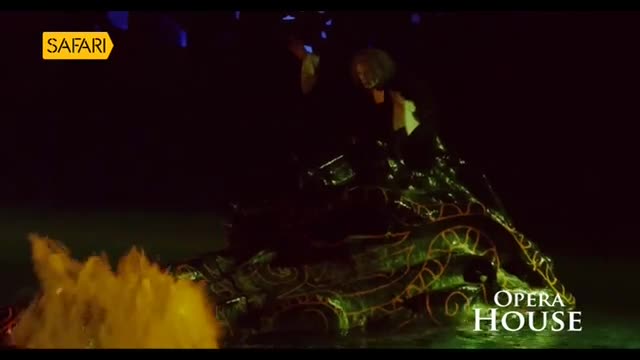 A performer, lit by a spotlight, stands on a glowing, ornate structure that resembles a dragon.  Yellow mist billows from the front as Safari TV presents the...