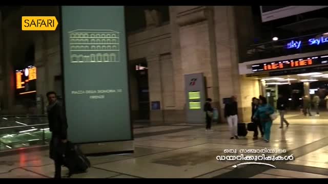 A man with a suitcase walks past a large sign in a European train station, likely for a segment on Safari TV. Other people are scattered around, some pulling...