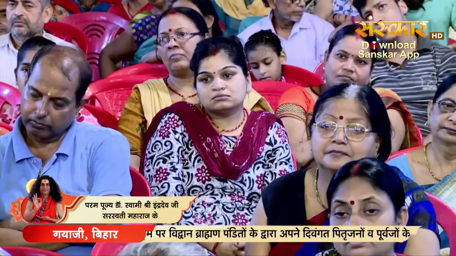 People are seated, listening attentively, some with their eyes closed. The event is being broadcast on Sanskar TV from Gaya, Bihar.