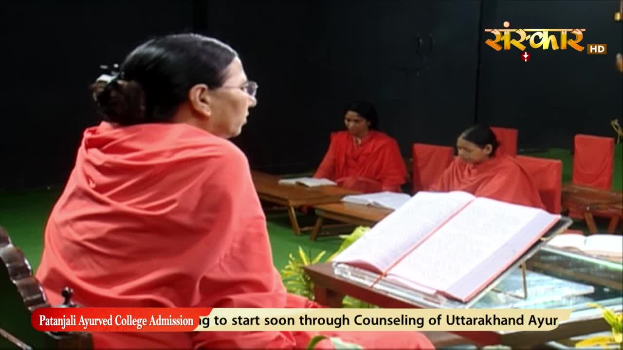A woman in a bright orange robe sits facing away, a large book open on a stand in front of her. Across the room, two other women in similar robes sit at tabl...