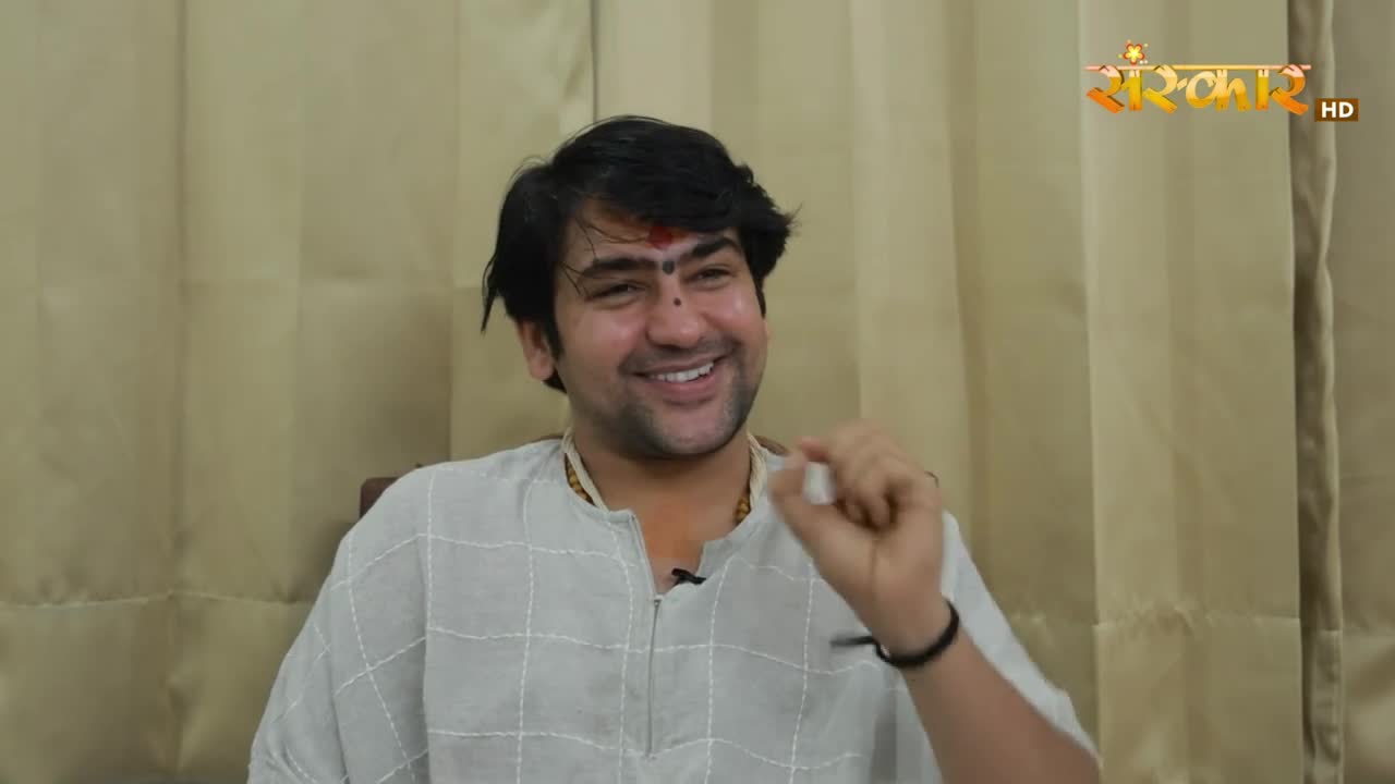 A man in a light-colored shirt, with a red mark on his forehead, smiles and gestures with his right hand. Behind him, the Sanskar HD logo is visible, suggest...