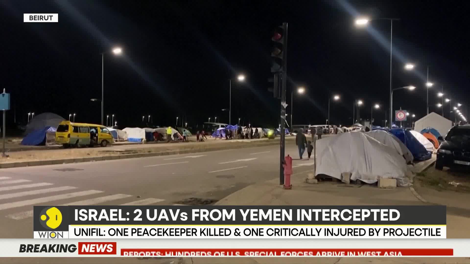 A line of tents stretches along the roadside at night, illuminated by streetlights. A yellow van is parked with its door open, and a few people walk by the t...