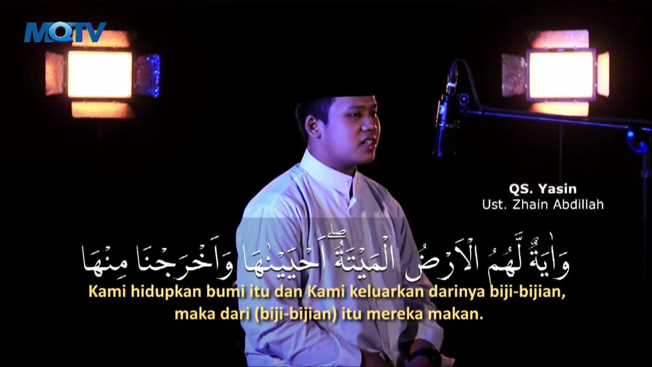 A man in a white shirt speaks into a microphone on a dark stage, with bright lights framing the scene. Arabic script and Indonesian translations appear below...