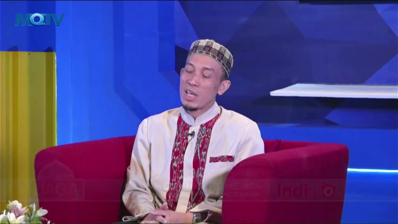 A man in a patterned cap and cream-colored shirt sits in a red armchair, speaking animatedly. The MQTV logo sits in the upper-left corner, and the background...