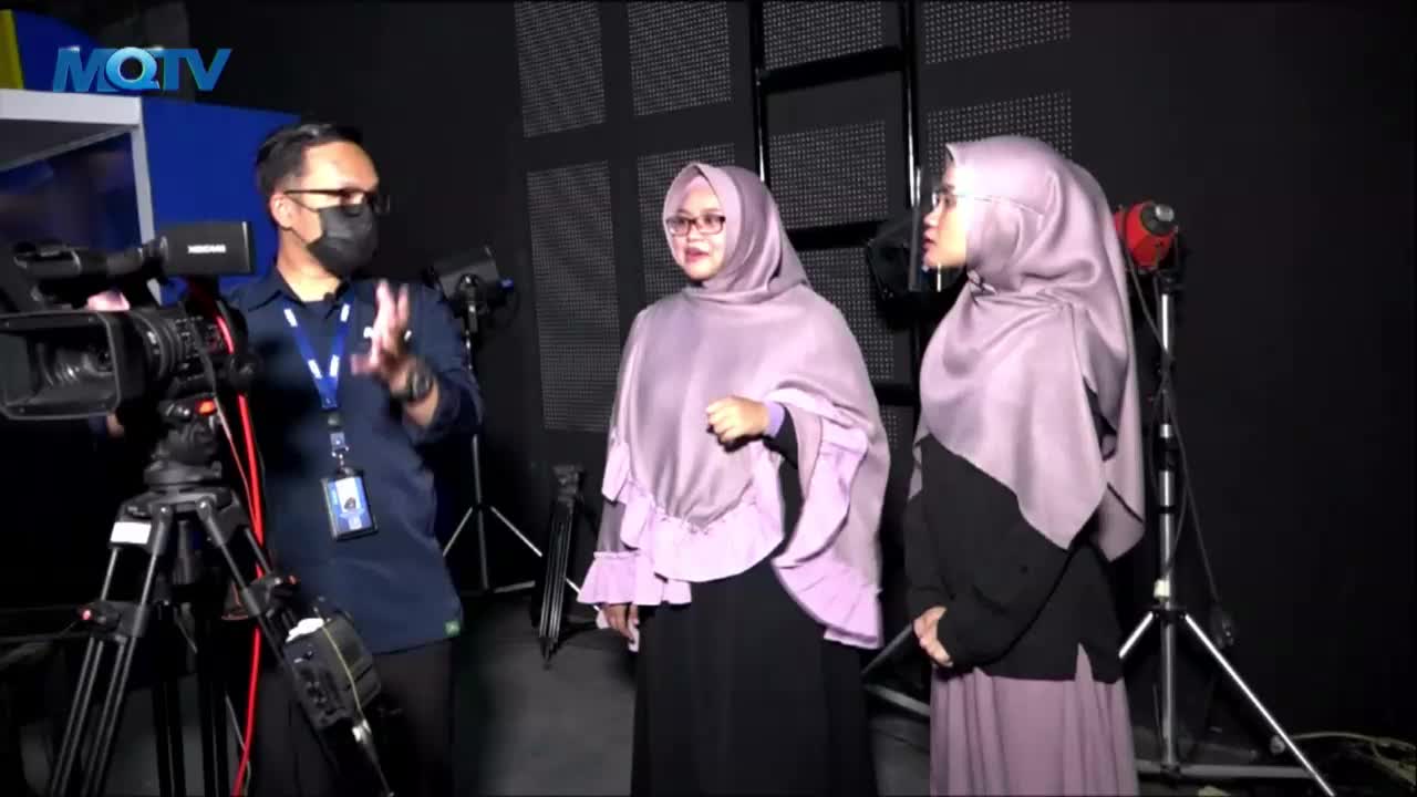 A man in a mask gestures with his hands, explaining something to two women wearing hijabs in an MQTV studio. The women stand still, listening, as a camera on...