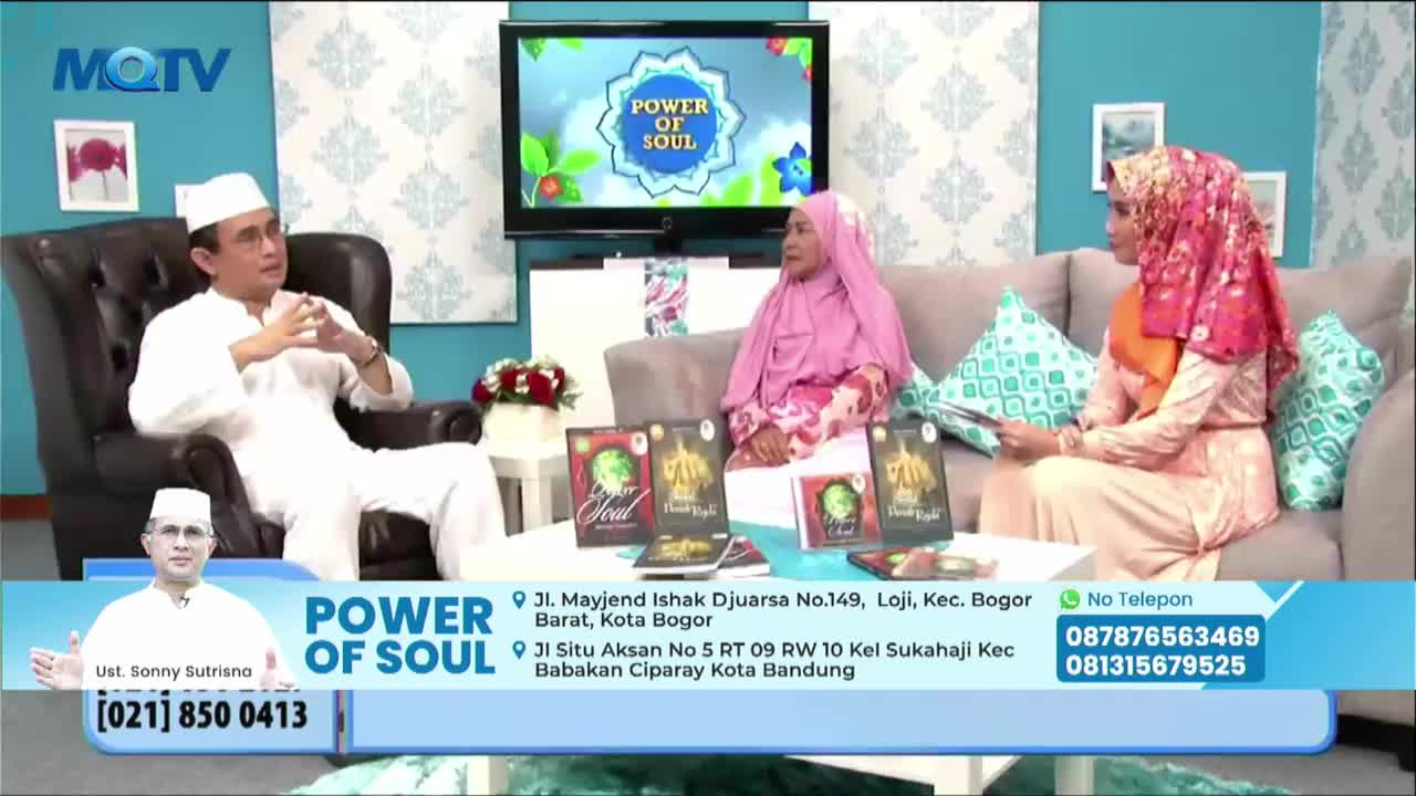 A man in white is speaking in a studio setting, gesturing with his hands. Several copies of a book are displayed in front of him and two other women.