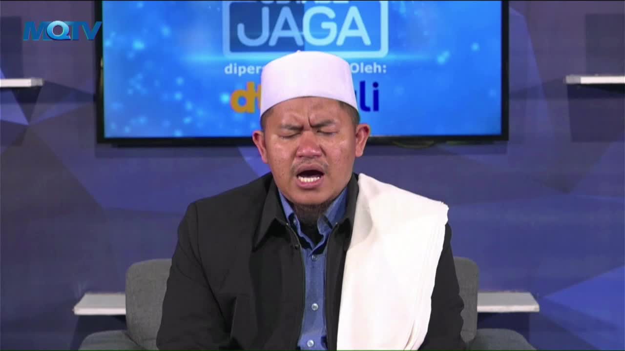 The man in the white cap and shawl is speaking, his mouth open as he looks upwards. Behind him, a screen displays the MQTV logo and the word "JAGA."