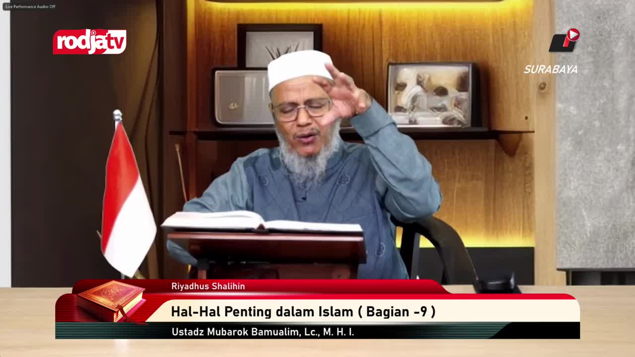 A man with a white cap and beard gestures with his right hand as he speaks, his left hand resting on an open book. The Indonesian flag stands to his left, an...