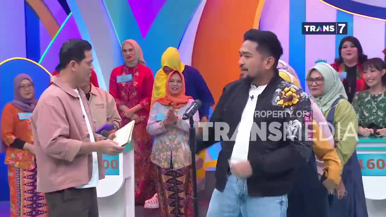 A man in a pink jacket holds a paper, facing another man in a black jacket with floral embroidery. Behind them, a crowd of people watch the proceedings on a ...