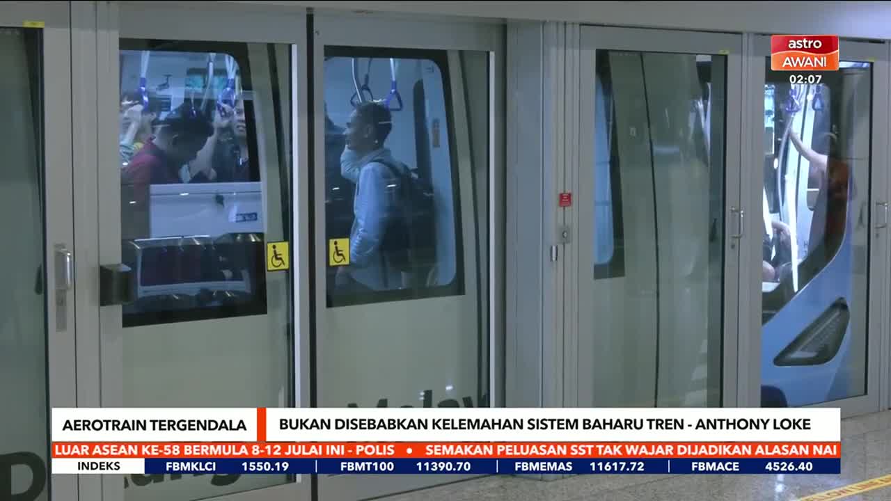 Astro Awani is reporting on a stalled aerotrain; passengers are visible inside the train's doors. A man with a backpack stands near the glass, and the doors ...