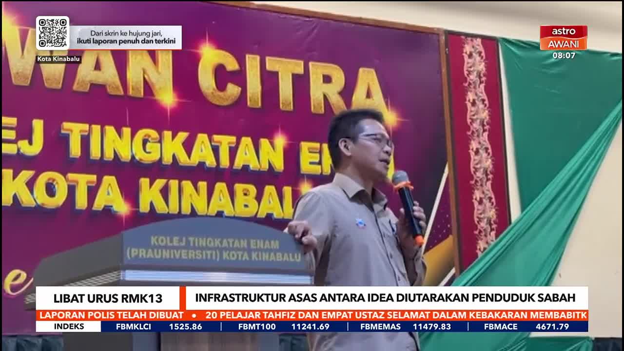 A man in glasses gestures with a microphone, speaking at a podium in front of a banner. The Astro Awani logo is visible on the right side of the screen.