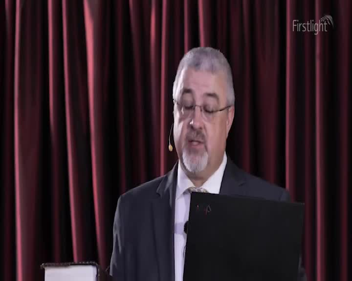 A man in a suit, wearing glasses and a lavalier microphone, is speaking, possibly reading from a laptop. Behind him, a burgundy curtain provides a backdrop f...