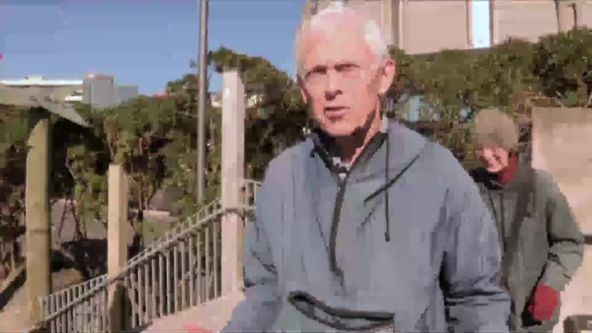 A man in a blue jacket walks towards me, his white hair catching the sun. Behind him, another person in a green jacket and beanie follows closely.