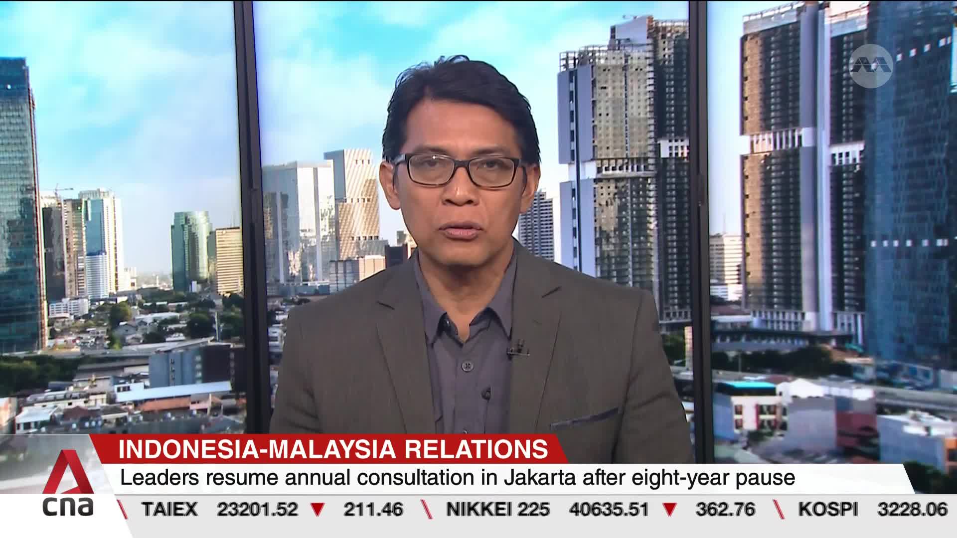 A man in glasses, wearing a blazer, is speaking directly to the camera. Behind him, a cityscape of tall buildings is visible through a large window, with the...