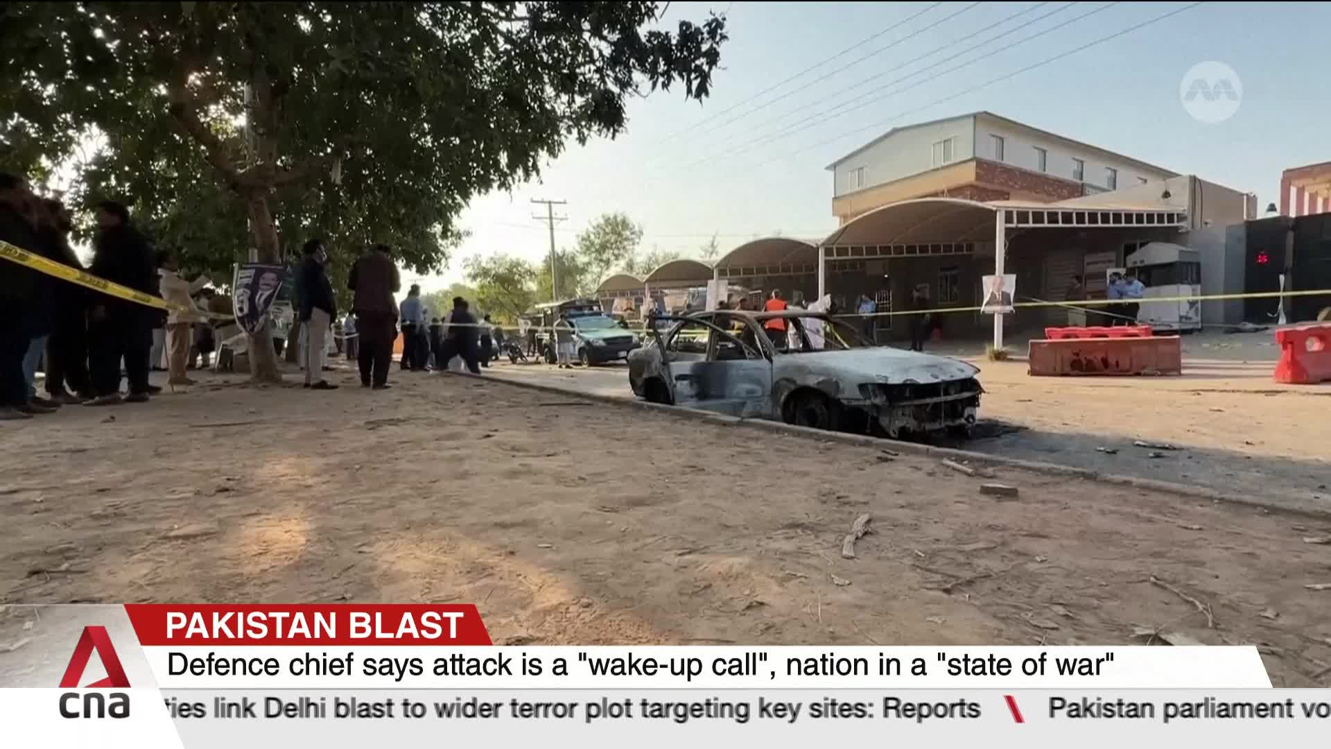 A burnt-out car sits on the side of the road, cordoned off with yellow tape. People stand and mill around, some looking towards the scene, as CNA Internation...