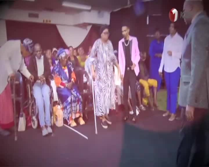 A woman in a zebra-striped dress, using a crutch, takes slow steps forward. She's being guided by a man in a pink jacket, while a crowd watches from their se...