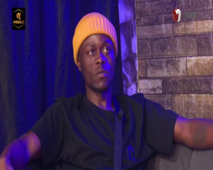 A man with a yellow beanie sits, looking intently toward the camera. He's on a set, with a 1KZN TV logo visible.