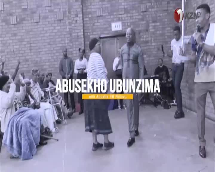 A man in a dark suit walks forward, while a woman in a striped shirt turns to face him.  People in the background raise their hands, and the 1KZN TV logo is ...