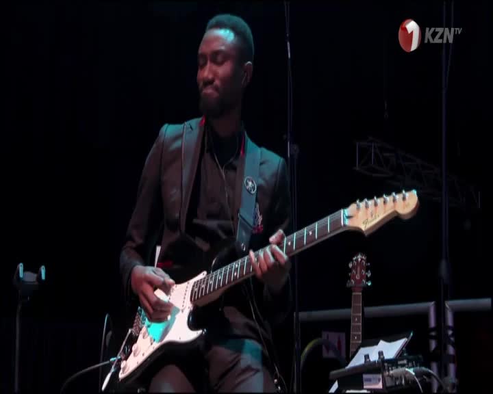 A musician, lit by a spotlight, strums a white electric guitar, his fingers dancing across the frets. 1KZN TV is broadcasting from South Africa, and the stag...