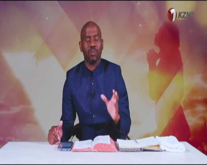 On 1KZN TV, a man in a blue patterned jacket gestures with his hands, speaking from behind a desk.  Open books and a phone sit before him, with a silhouette ...