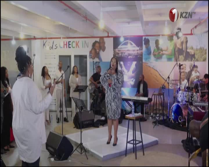 A woman in a zebra-print dress sings into a microphone on a small stage. Behind her, a band and backing vocalists accompany the performance, which is likely ...