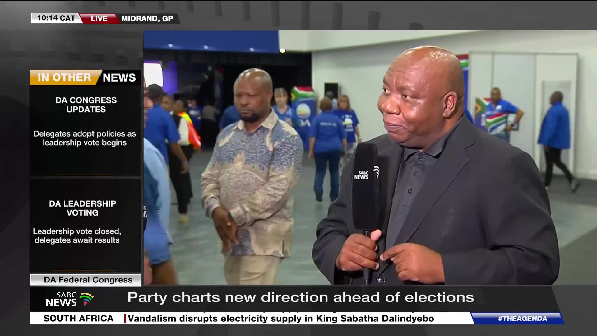 A man in a black suit holds an SABC News microphone, speaking to the camera. Behind him, people in blue shirts move through a hall, some carrying South Afric...