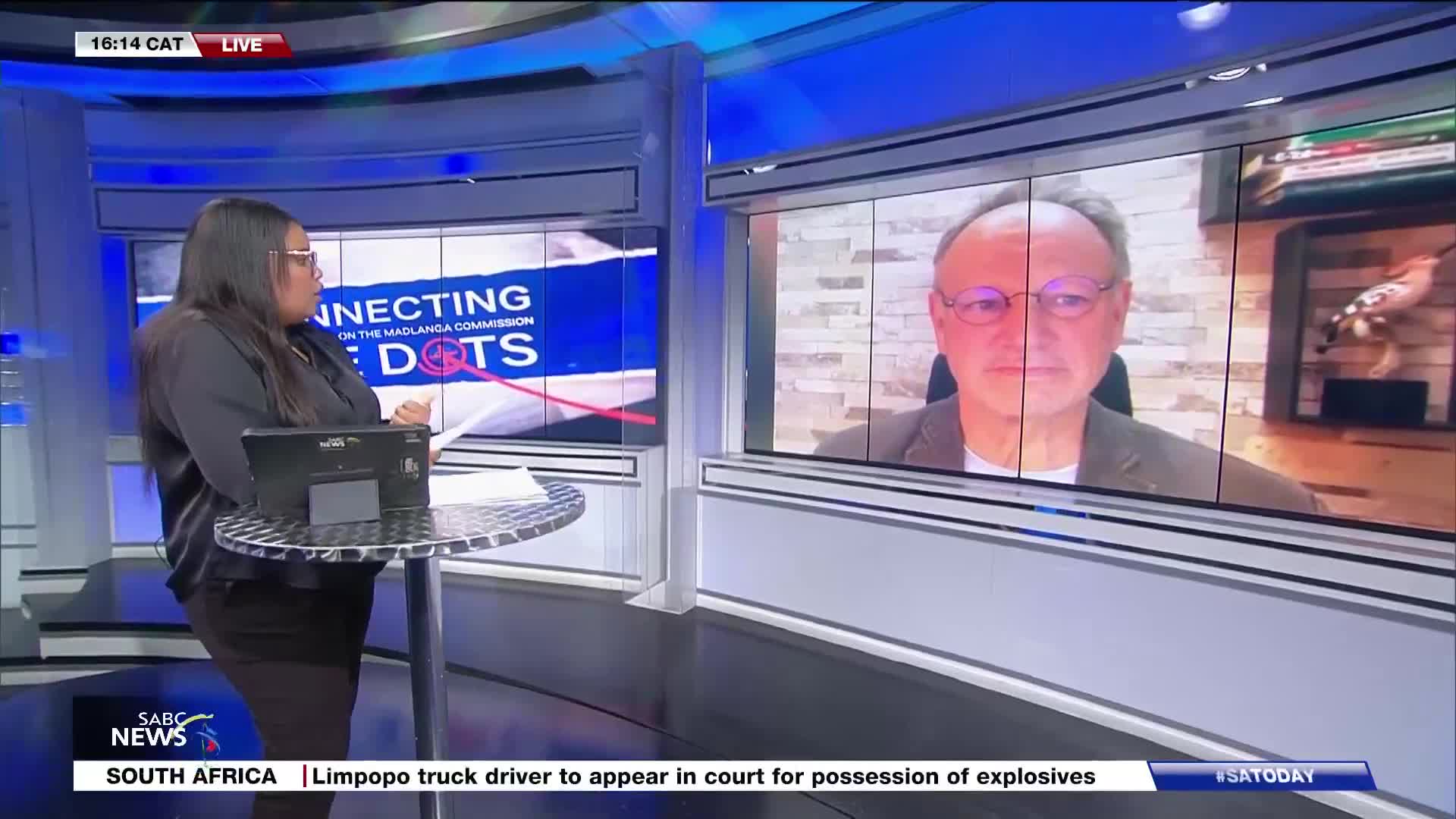A woman in a dark shirt stands at a metal table, looking at papers while a man in glasses appears on a large screen behind her. The SABC News logo is visible...