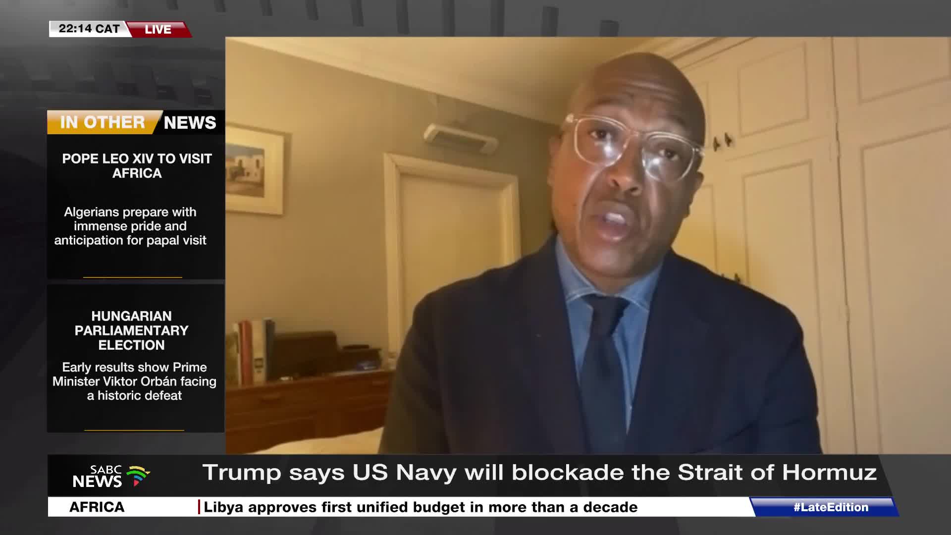 A man in a suit and tie speaks directly to the camera, his glasses reflecting the light. On the left, SABC News displays headlines about a papal visit to Afr...