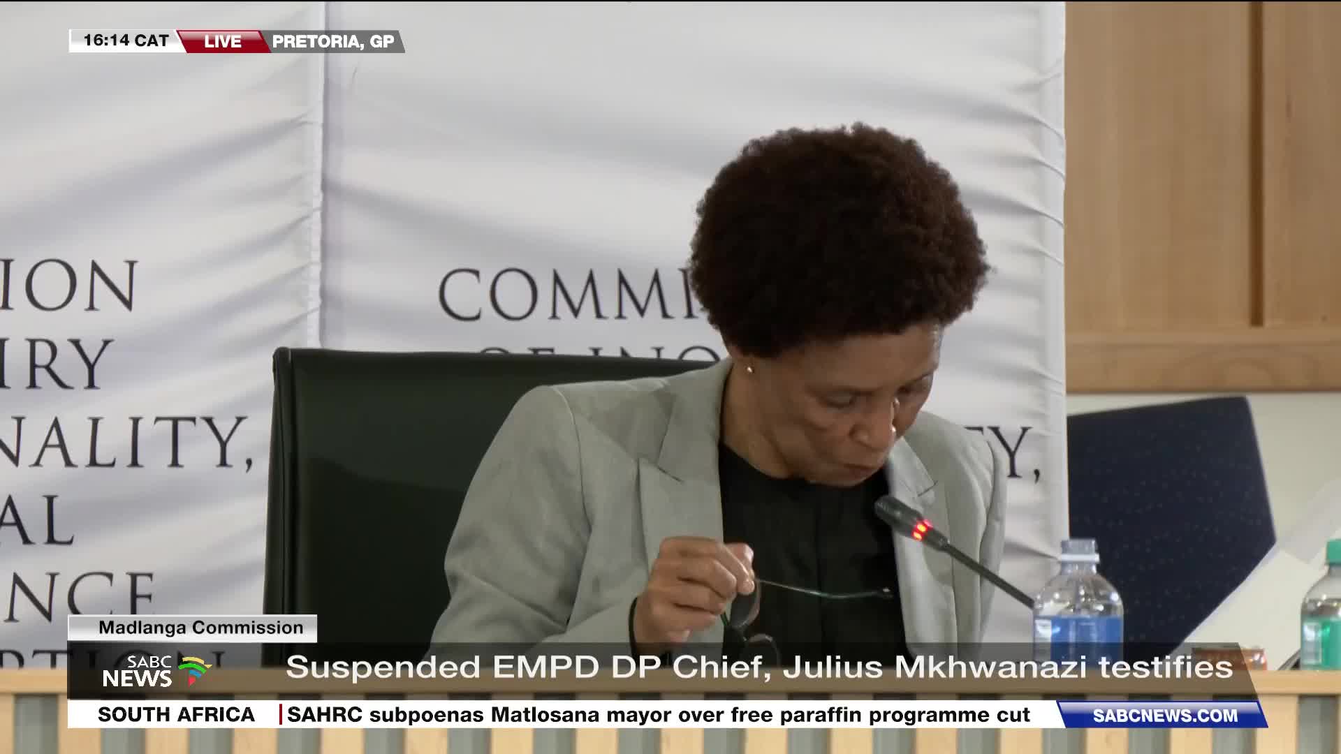 A woman in a light grey blazer leans forward, holding her glasses as she looks down. The SABC News banner at the bottom of the screen indicates this is a liv...