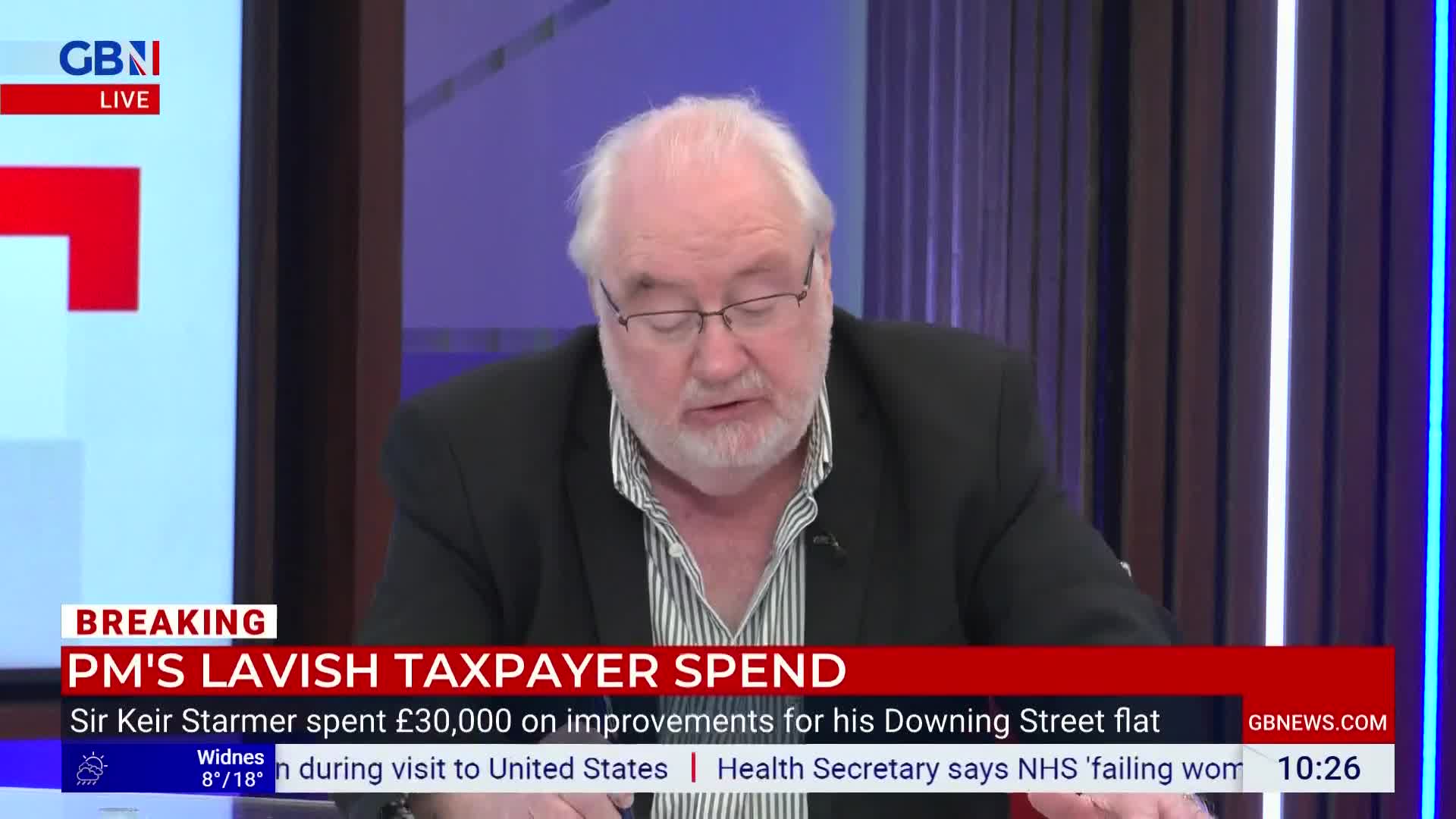 A man with white hair and glasses speaks into a microphone, his gaze directed downwards. Behind him, a screen displays "GBN LIVE" and a breaking news headlin...