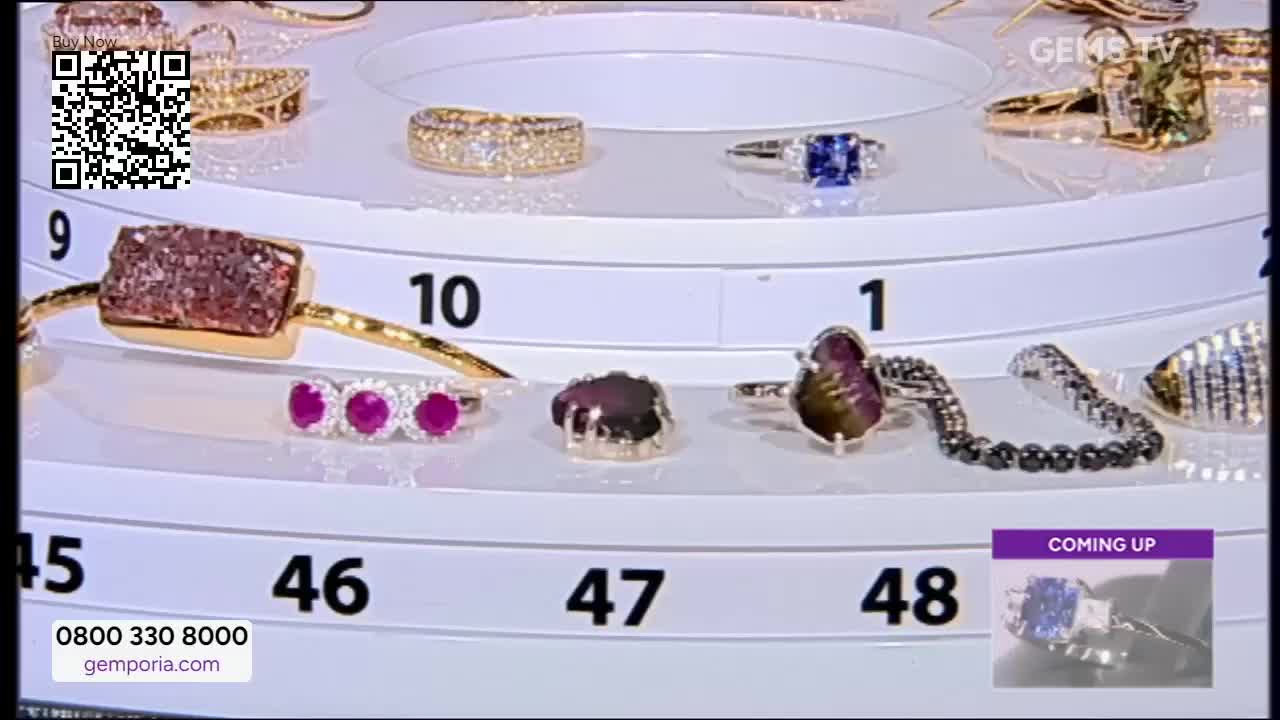 A rotating display of jewelry is set up, with each piece numbered and lit. The Gems TV logo is visible, and a "Coming Up" preview of a ring with a blue stone...