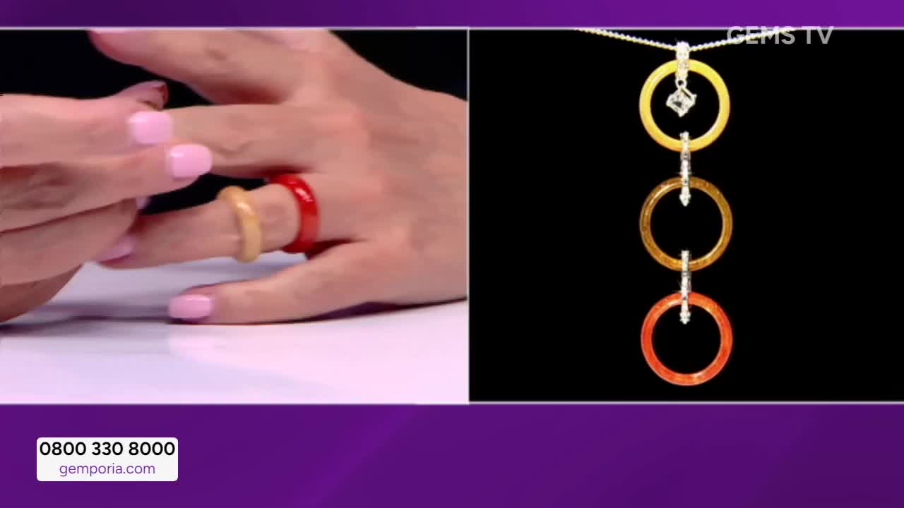 A hand on Gems TV displays two rings, one red and one yellow, on a finger. To the right, a necklace with three matching colored rings hangs against a black b...