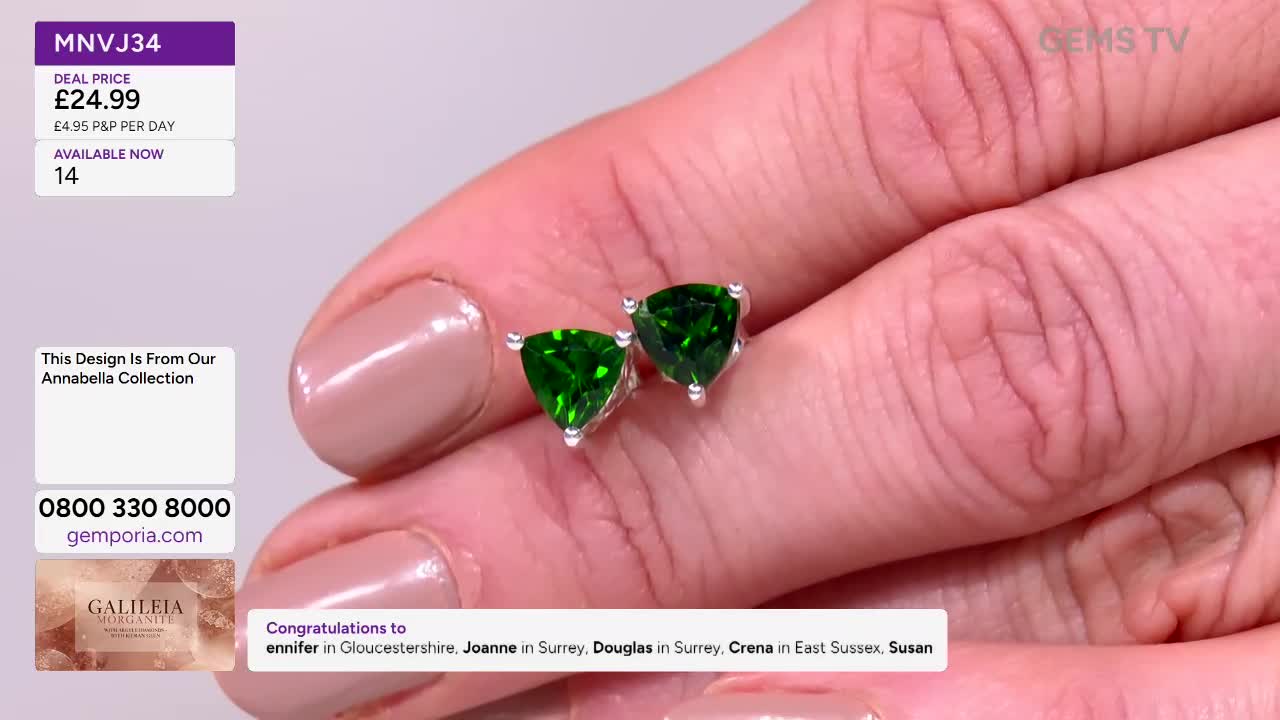 A pair of emerald-green, triangular earrings are held up for the camera on Gems TV. The presenter's fingers, with neatly manicured nails, cradle the jewelry.
