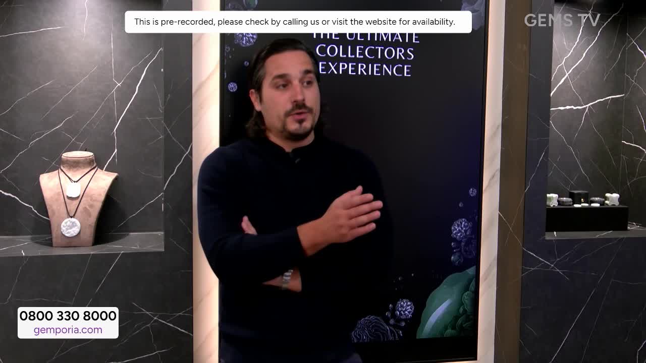 A man in a dark sweater gestures while speaking on a Gems TV set. Behind him, a display shows jewelry and a screen promoting "The Ultimate Collectors Experie...