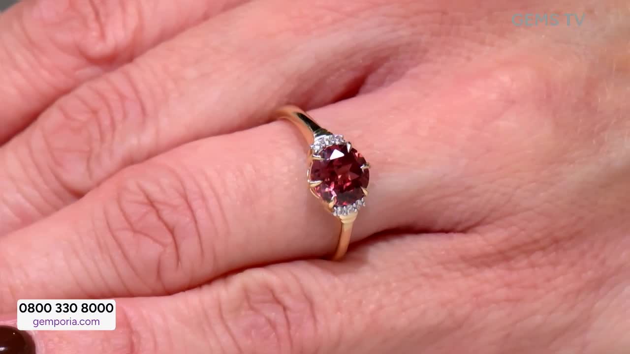 On Gems TV, a hand displays a gold ring with a large, oval, red gemstone flanked by smaller white stones. The ring is held up, catching the light as if ready...