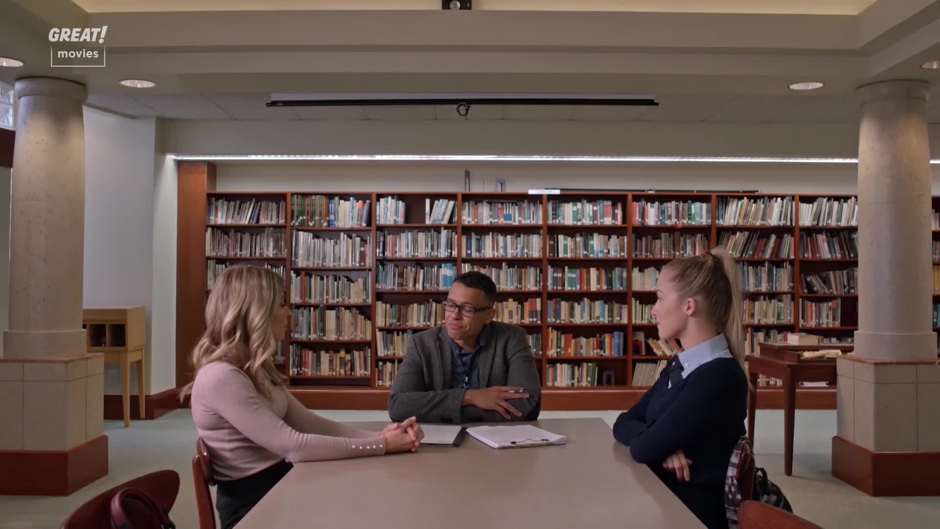 Three people sit around a table in a library, facing each other. The man in the middle is speaking, while the two women listen intently, their hands clasped ...
