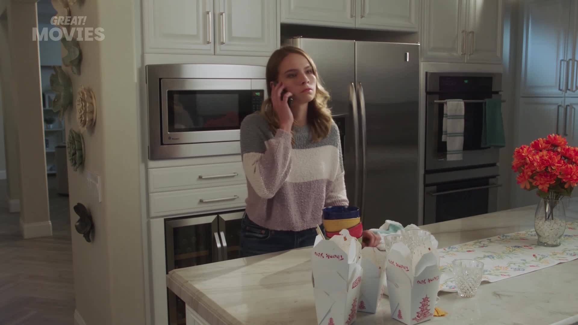 A young woman stands by the counter, talking on her phone, with several takeaway containers lined up. A vase of bright orange flowers sits on the counter to ...