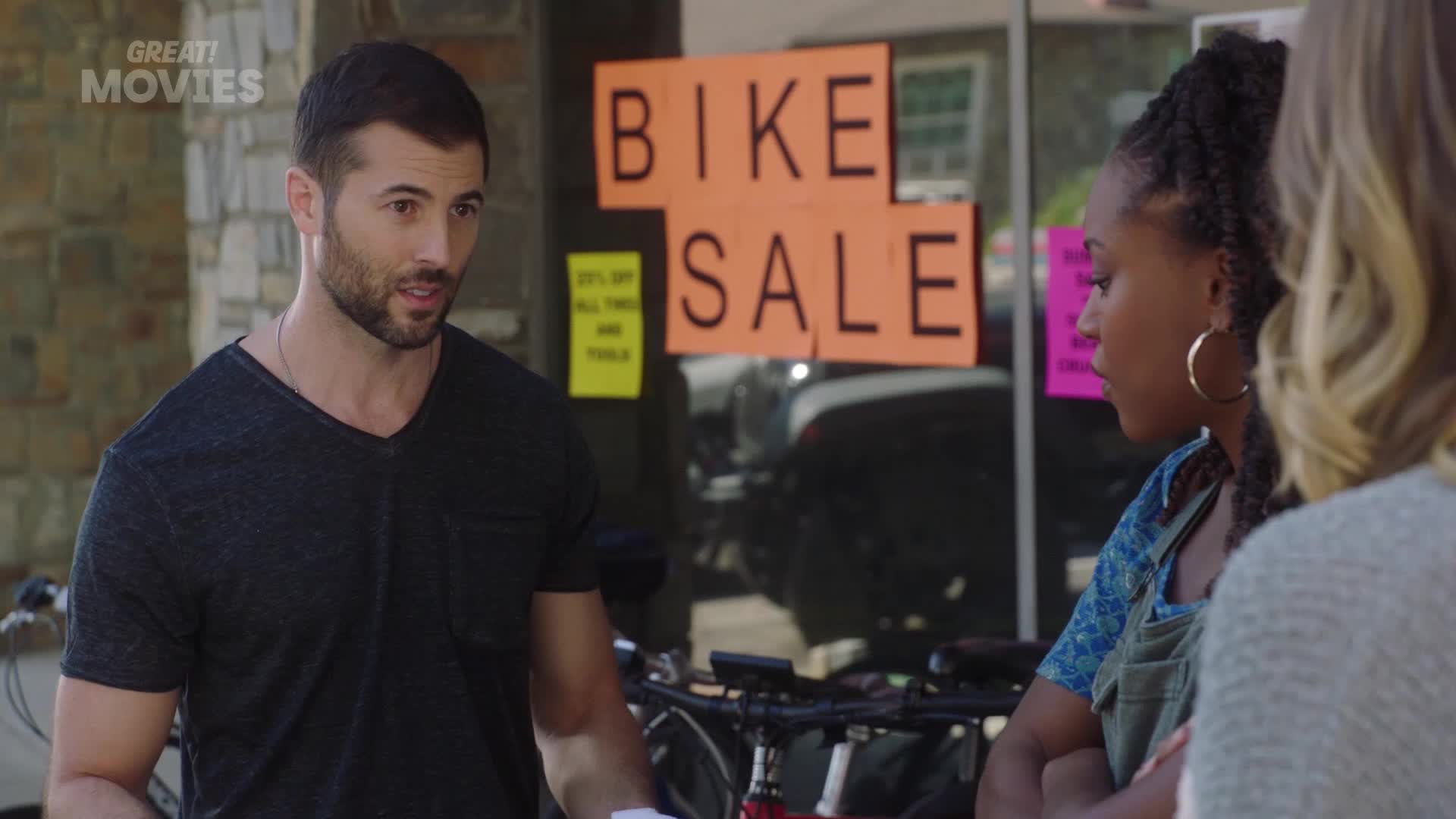 A man in a dark t-shirt gestures as he speaks to two women outside a shop with a "BIKE SALE" sign in the window. The women, one with large hoop earrings, lis...