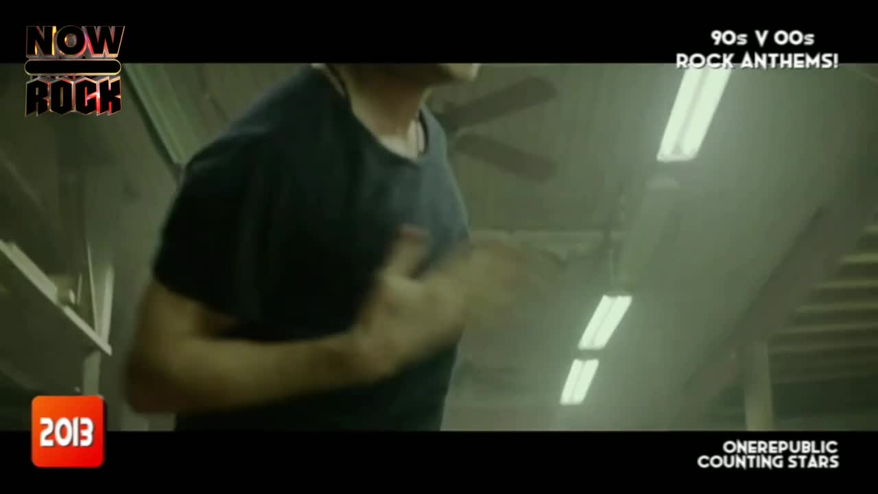 A man in a dark t-shirt is clapping, his arms moving quickly. The scene is dimly lit, with a ceiling fan and fluorescent lights visible in the background.