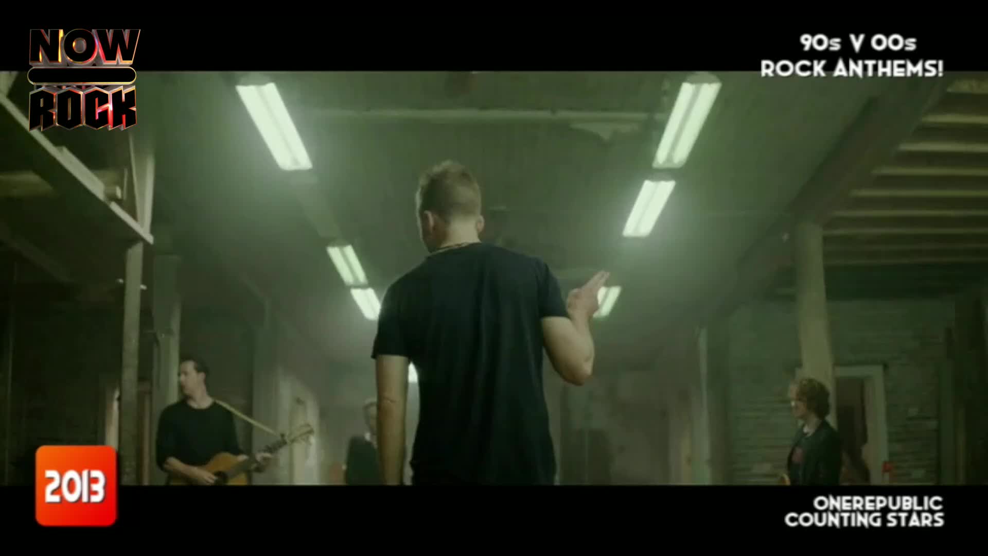 A man in a black t-shirt gestures as he walks down a long, dimly lit corridor. Behind him, a band plays, illuminated by the overhead fluorescent lights.