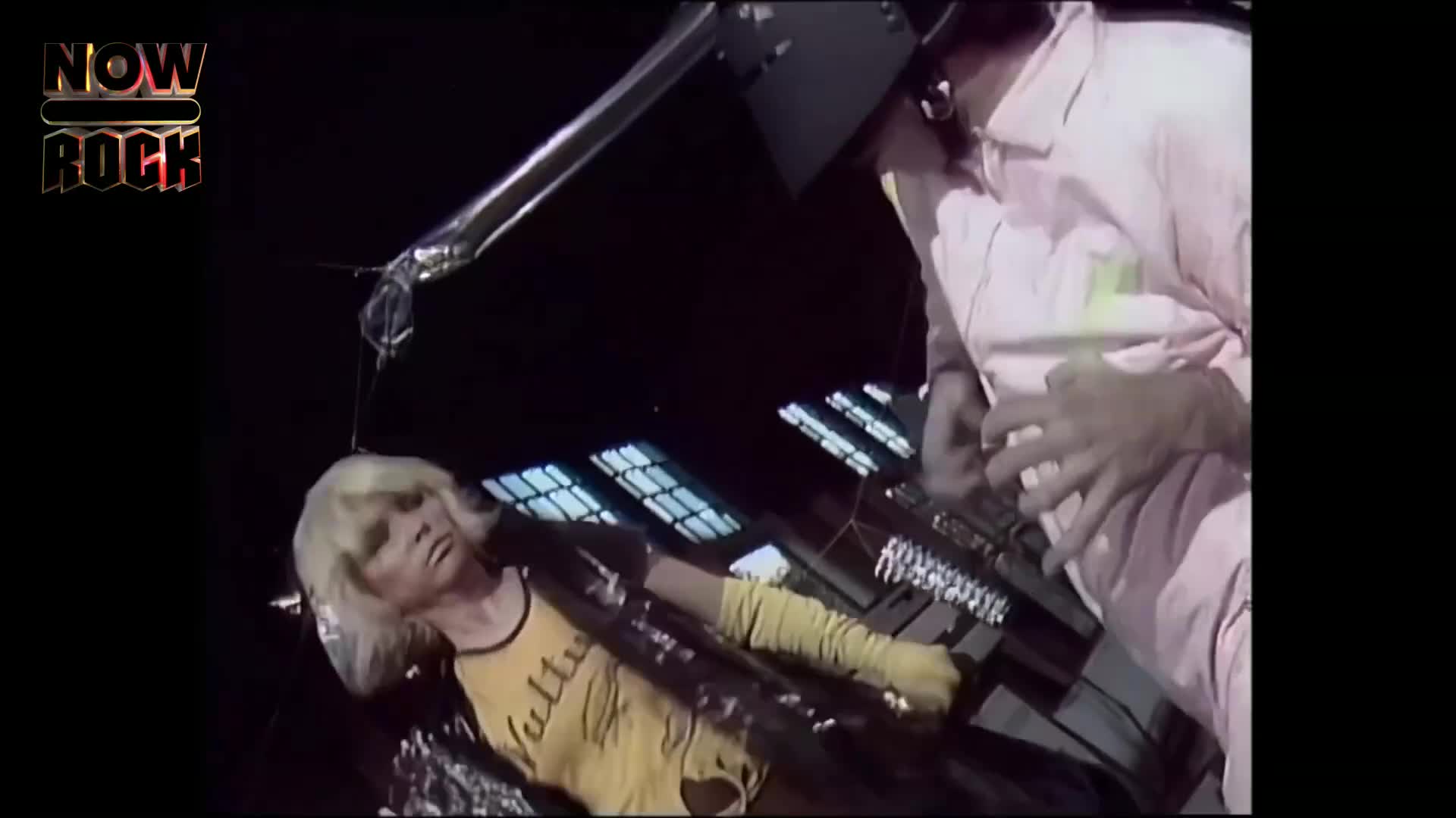 A woman in a yellow shirt and long gloves is lying down, playing a keyboard. A man in a pink suit is standing over her, working with a different keyboard.